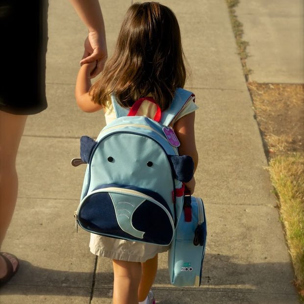 Una estudiante pequeña con mochila de elefante toma la mano de un adulto camino a la escuela